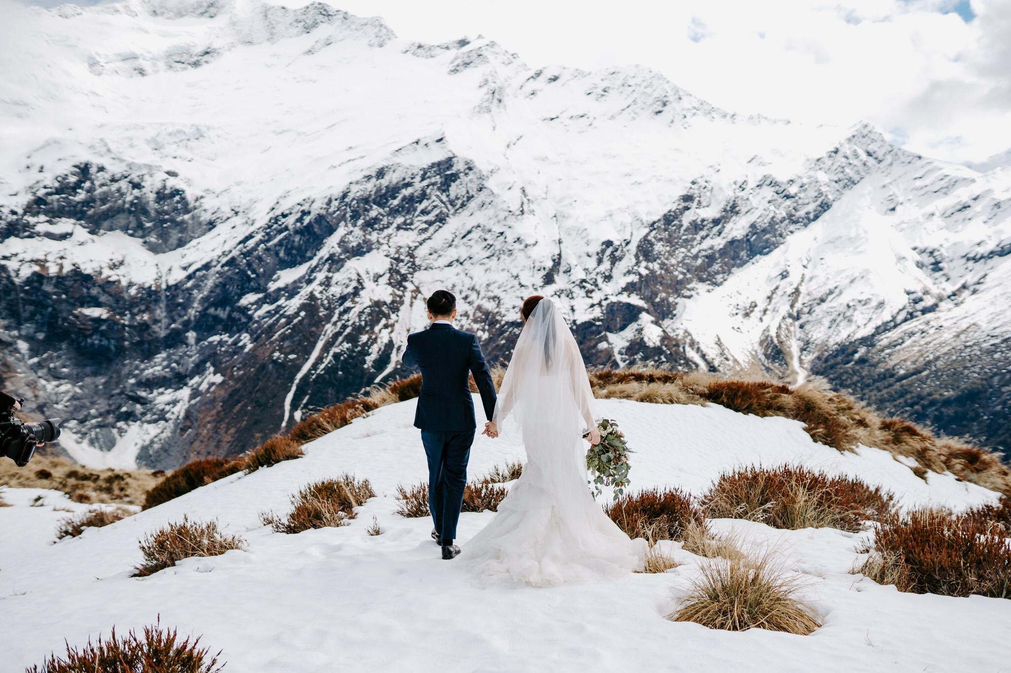 A picture of a newly married couple standing on a rocky mountain peak, surrounded by the stunning natural scenery of New Zealand. The couple is dressed in their wedding attire, with the bride wearing a white wedding dress and the groom wearing a black suit. They are holding hands, taking in the breathtaking views of the mountains around them.