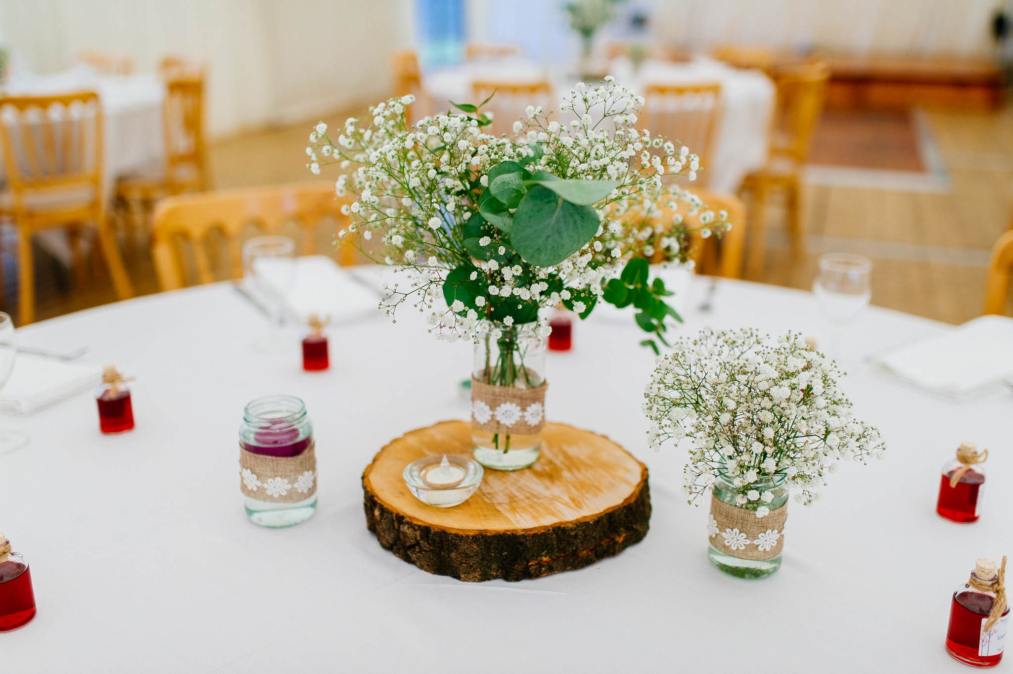 A captivating boho-inspired centrepiece on a wedding table. A rustic wood log slice serves as the base, adorned with a delicate vase filled with whimsical boho flowers. The vibrant blooms feature a mix of wildflowers, feathers, and lush greenery, creating a dreamy and enchanting display.