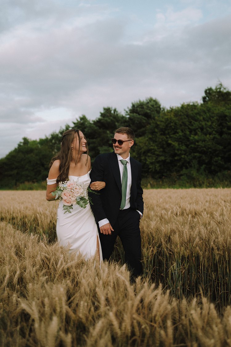 A blissful newly married couple strolls hand in hand through a lush crop field, their faces radiating pure joy and love, bathed in the golden light of their wedding day.